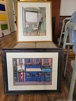 Two framed art prints stacked vertically, one with interior scene, one storefront scene