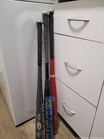 Three metal baseball bats leaning against kitchen cabinets and a refrigerator on a wooden floor.