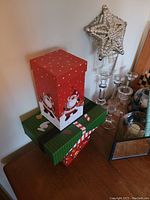 View of two holiday themed gift boxes with Santa and candy cane patterns placed on a table corner. Adjacent to glass candle holders and decorative items.