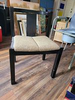 Photo showing front and side view of black wooden chair with beige upholstered seat on wooden floor with artworks in the background.