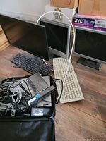 Two computer monitors (one black framed larger type, one gray framed smaller type), two keyboards, various cables including VGA cable, and a black HP portable device laid out on a wood floor, several cardboard boxes in background.