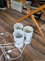 Set of three white cylindrical floor lamps shown assembled with cords extended, placed on hardwood floor under furniture.