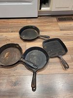 Four cast iron pans on wooden floor: three round frying pans and one square grill pan, all heavily used and needing cleaning.