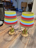 Pair of table lamps with colorful striped fabric shades and wooden bases with frog decorations, placed on wood floor near furniture.