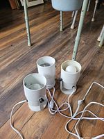 Three small white cylindrical electric accent lights on wood flooring, two without bulbs and one with a white bulb installed, showing on/off switches and cords tangled on the floor.