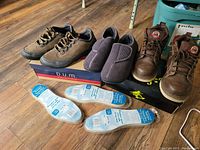 Three pairs of men's shoes displayed on boxes on wooden floor: casual sneakers, slippers, and work boots plus three pairs of insoles laid out in front.