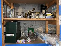 Wide view of multiple shelves containing various candle holders made of glass, brass, ceramic, and metal. Visible are stacked brass candlesticks and several glass holders, candles, and ceramic flower-shaped candle holders.