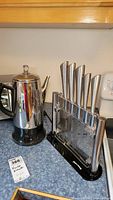 Overview photo of stainless steel Cuisinart electric coffee percolator next to clear acrylic knife holder containing six stainless steel knives with brushed handles on blue countertop.