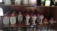 Shelf displaying multiple tall cut-to-clear crystal glasses with cranberry rims, stemmed goblets with cranberry rims, and a cream and sugar set.