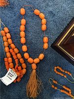 Four strings of orange Greek Koloboli beads laid out on a blue carpet alongside a picture frame. Strings have tassels attached in orange and yellow hues.