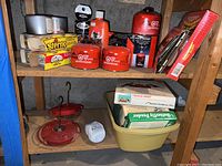 Photo of wooden shelf holding Sterno cooking fuel cans and containers, red hummingbird feeder parts, boxed fuel gels, and other related items.