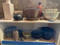 Overview of multiple ceramic pottery items including plates, bowls, vases, and cups on a white shelf with a concrete wall background.