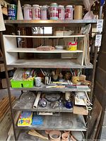 Photo of shelving unit with pottery supplies including glaze jars, pottery tools, brushes, sponges, ceramic chucks, and other miscellaneous pottery accessories.