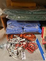 Stack of green and blue tarps wrapped in plastic, with coiled orange and white extension cords below and hanging work light attached to an orange cord.