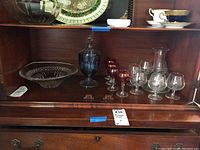 Overall view showing Waterford crystal bowl, lidded blue glass bon bon jar, colored and clear stemware glasses, and clear carafe arranged on wood shelf.