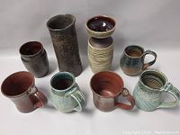 Front view of eight signed pottery pieces including vases and mugs placed on a grey surface showing colors and shapes.