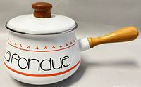 Side view of vintage enamel fondue pot displaying wooden handle and printed 'La Fondue' on white body.