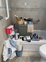 Photo showing large woven laundry basket with lid, various bath and hair products on it, blood pressure monitor box, soap, scale, towels, and plastic cups in a bathroom setting.