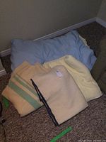 Blue comforter on floor next to three folded blankets: beige striped wool blanket, cream wool blanket with blue edging, and yellow throw blanket.