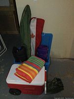 The full camping lot showing the cooler, camping chairs in cases, rug, sleeping bag, storage bin, and portable table.