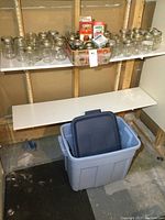 Shelf with about 30 glass canning jars of various sizes, mostly with metal lids, some without lids. Two cartons of Certo pectin mix visible in the background. Blue plastic storage bin with lid on floor below shelf.