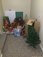 Photo showing two artificial Christmas trees, multiple wreaths, gift bags, gift wrap, ribbons, and other decorations arranged near carpeted steps.