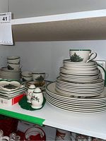 Full view of stacked plates, cups, saucers, salt and pepper shakers, and other accompanying pieces all showing a consistent green rim and Christmas tree holiday pattern.