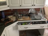 Wide view of kitchen counter and open cupboard with metal nesting bowls, glass dishes, wire basket, cookbook, cutting boards, and cutlery drawer open showing various metal cutlery.