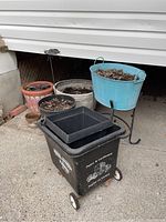 All items in the lot including the black wheeled cart, smaller planters and large blue tin planter on stand visible under house eave.