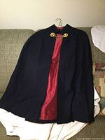 Black nurse cape hanging on a wire hanger, showing maroon lining and gold chain clasp on the collar.