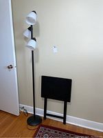 Full view of black metal floor lamp with three white plastic shades alongside black wooden TV folding table folded and leaning against the wall on hardwood flooring.