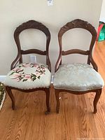 Two matching carved wood parlour chairs with different upholstered seats next to each other, showing front view, highlighting carved floral wood details on back and upholstery difference.