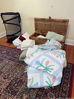 Entire lot showing quilt, bedding, wicker basket, laundry bag and linens on floor and rug.