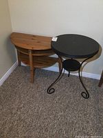 Both tables pictured together in a corner on carpet, showing the wood D-shaped end table behind the black metal round table.