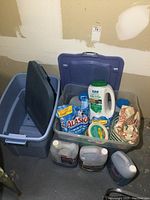 Two large plastic totes, one empty with lid, the other filled with bags of ice melter, jugs of windshield washer fluid and containers.