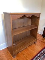 Photo of a medium brown wooden bookcase with decorative scalloped edges, two fixed shelves, and vertical slat back panel, showing overall condition including minor wear.