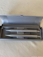 Two silver ballpoint pens inside the original silver-colored metal gift box with foam insert, showing pens in place, unopened and unused.