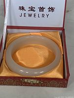 White jade bangle bracelet resting inside a red and gold decorated jewelry box with yellow satin interior, showing overall lot presentation.