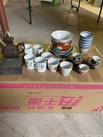 Photo showing multiple porcelain cups, bowls, a metal box and Zisha teapot arranged on a cardboard box.