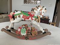 Side view of the white porcelain rocking horse music box decorated for Christmas with detailed painted accents including red bows, green saddle, gold bells, and holly leaves. It rests on a brown wooden rocker base with gifts and toys underneath.