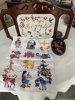 Wide shot showing three embroidery decoration pieces: wooden framed mirror with embroidery, oval embroidery hanging, and embroidered cloth with figures spread on the table.