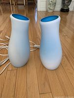 Two white curvy table lamps with matte finish and blue interiors standing next to each other on a wooden floor, showing their shapes and colors.