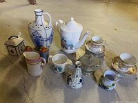 Front angled view of all porcelain items grouped including teapot, vase, multiple mugs, cups, saucers, covered jar, creamer, small pitcher, and glass dish with lid