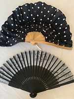 Photo showing two folding handheld fans, one black with white polka dots and bamboo ribs, the other gray with black floral patterns and black ribs.