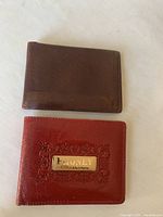 Two leather wallets displayed flat on a white surface, top wallet brown smooth leather bi-fold, bottom wallet red textured leather with metal logo plaque.