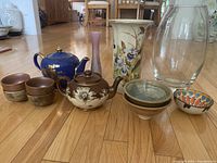 Photo showing two teapots, multiple bowls, and three vases arranged on a wooden floor.