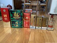 Photo showing full lineup of seven vintage Chinese liquor bottles with original boxes on wooden floor with chairs in background.