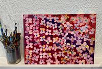 Front view of abstract floral oil painting showing predominantly pink and lavender flowers against a red background, with paintbrushes in a container beside it.