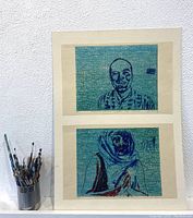 Photo of two stacked poster art prints featuring blue textured backgrounds and hand-drawn portraits, with a container of paintbrushes beside them.