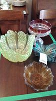 Four vintage art glass pieces on wooden table including a green hobnail bowl, red-rimmed compote, blue-green leaf dish, and amber scalloped bowl.